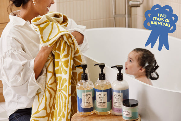 Mama bathing her toddler in bathtub with blue ribbon on upper right corner that reads "Two Years of Bathtime"