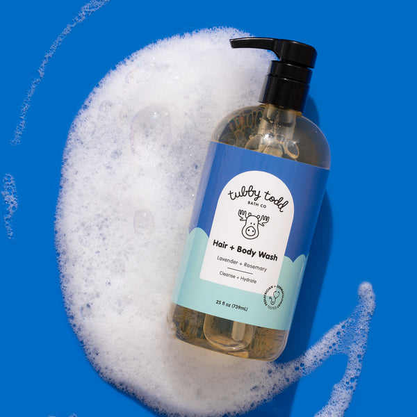 A bottle of Tubby Todd Hair + Body Wash rests on a vivid blue background surrounded by a large patch of white soap foam. The bottle is positioned diagonally and features a blue-and-mint label with the Tubby Todd logo and the scent “Lavender + Rosemary.” The black pump top contrasts sharply against the bright backdrop.
The bubbly foam creates a playful, fresh, and clean aesthetic while emphasizing the product’s lathering quality. Strong lighting and bold color contrast give the image a polished, modern prod