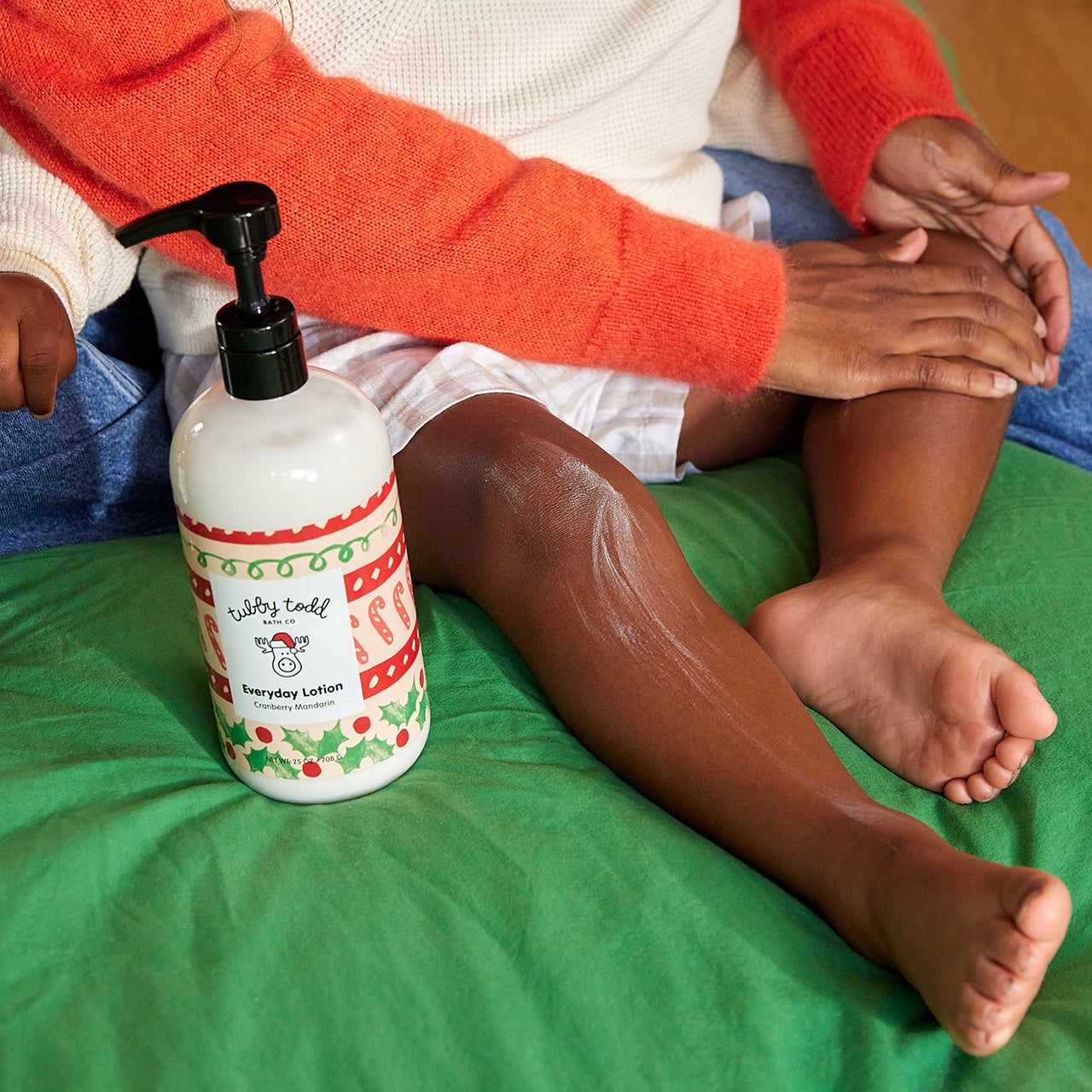 Mom applying the 25oz Cranberry Mandarin Everyday Lotion on her boy's leg on the bed.