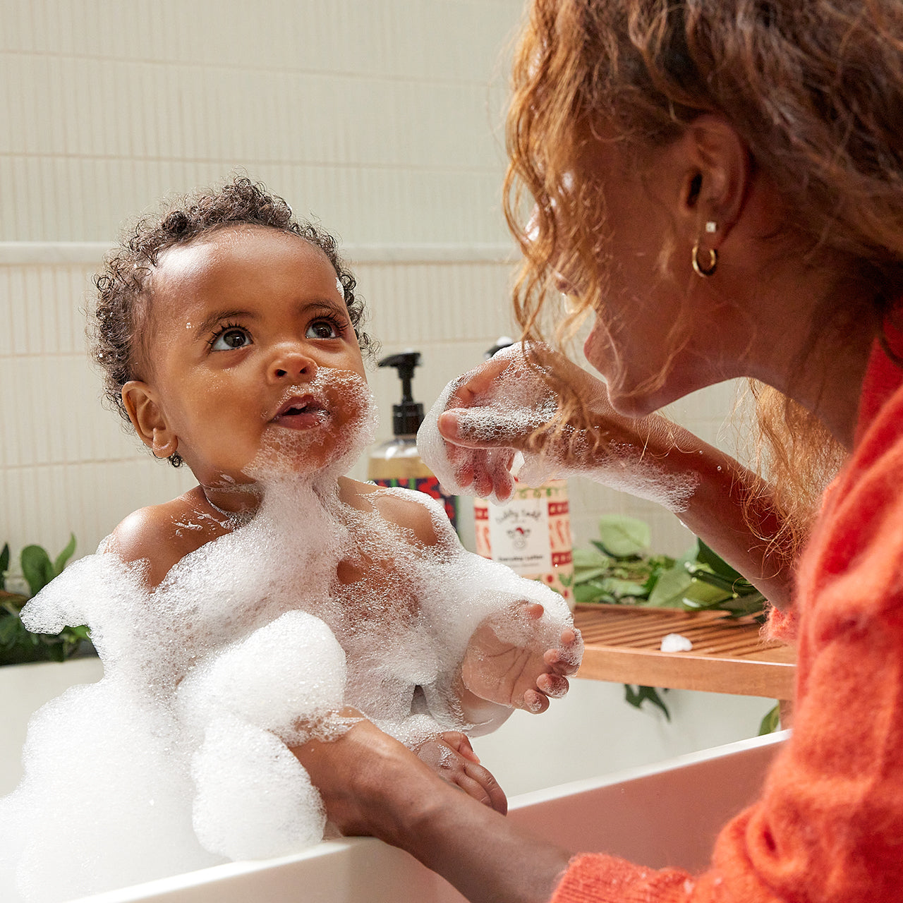 A baby covered in bubbles is taking a bath with his mom in the bathtub.