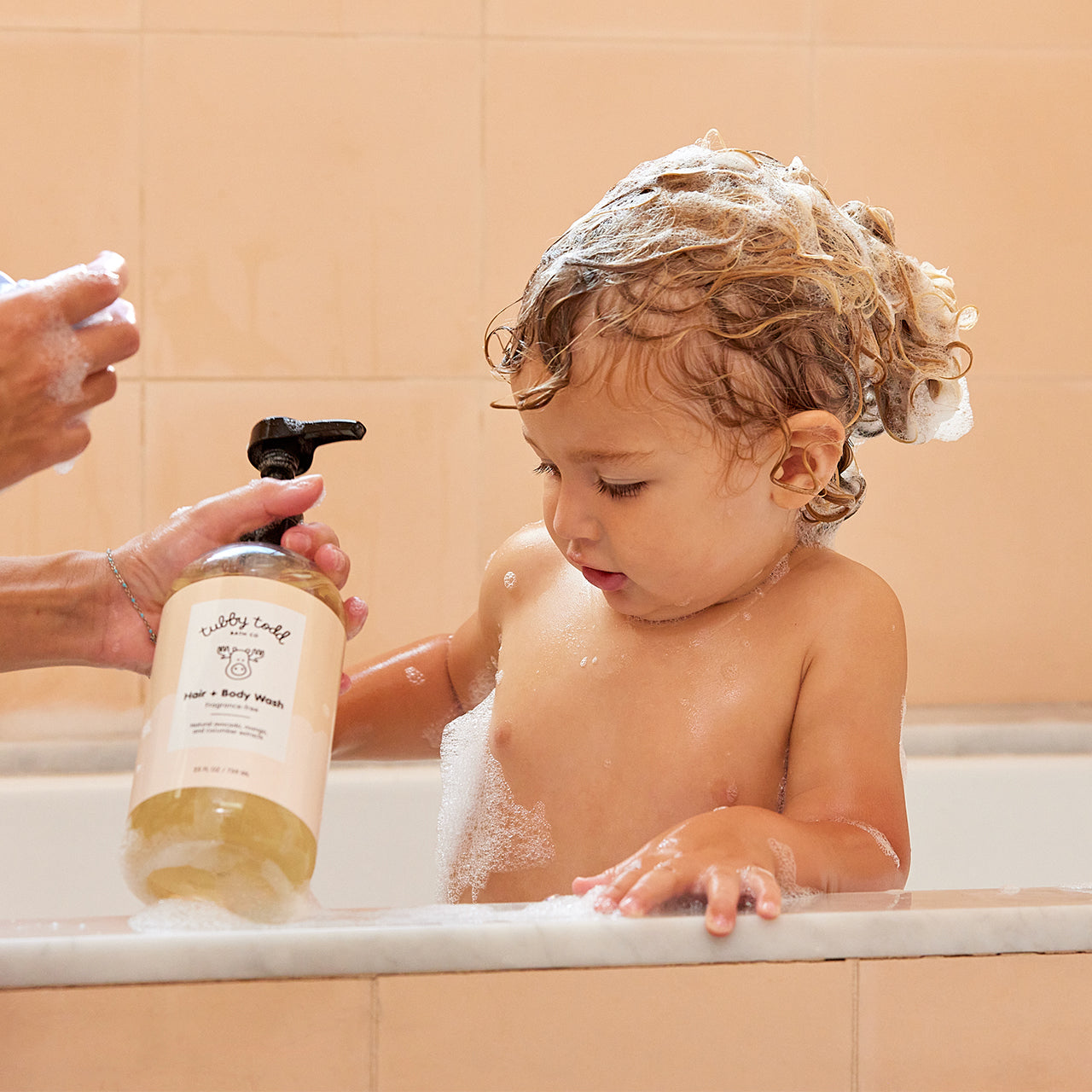 Mom holding the Fragrance-free Hair Body Wash bottle and her baby boy standing in the bathtub with bubbles on the hair.
