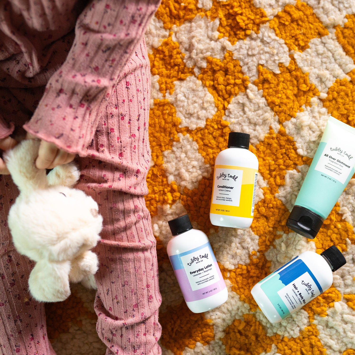 Travel Kit products lying on the colorful carpet and a girl sitting next to them with her rabbit doll.