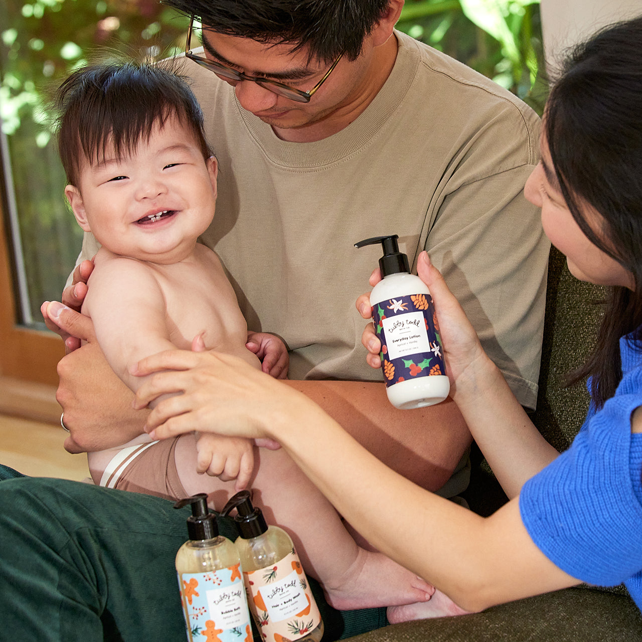 Baby boy smiling sitting on his dad's leg and his mom applying Tubby Evergreen's Everyday Lotion on his son's arm.