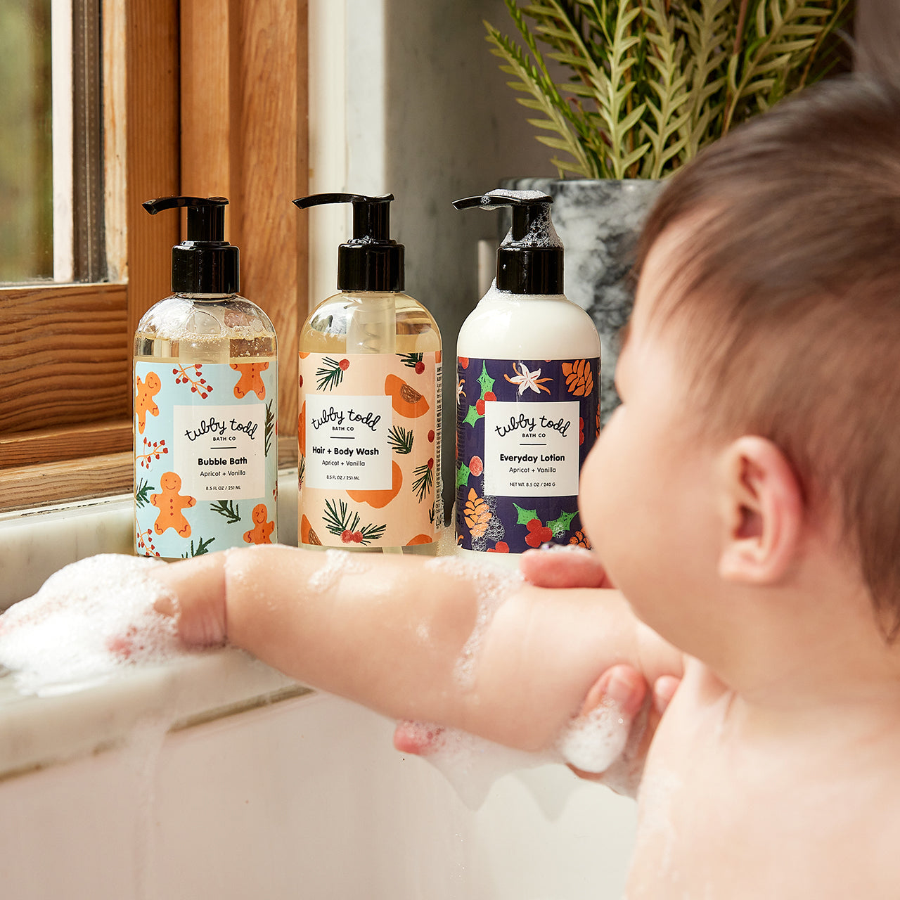 Baby boy with bubbles on his arms looking at the bottle of Tubby Evergreen bottles in the sink.