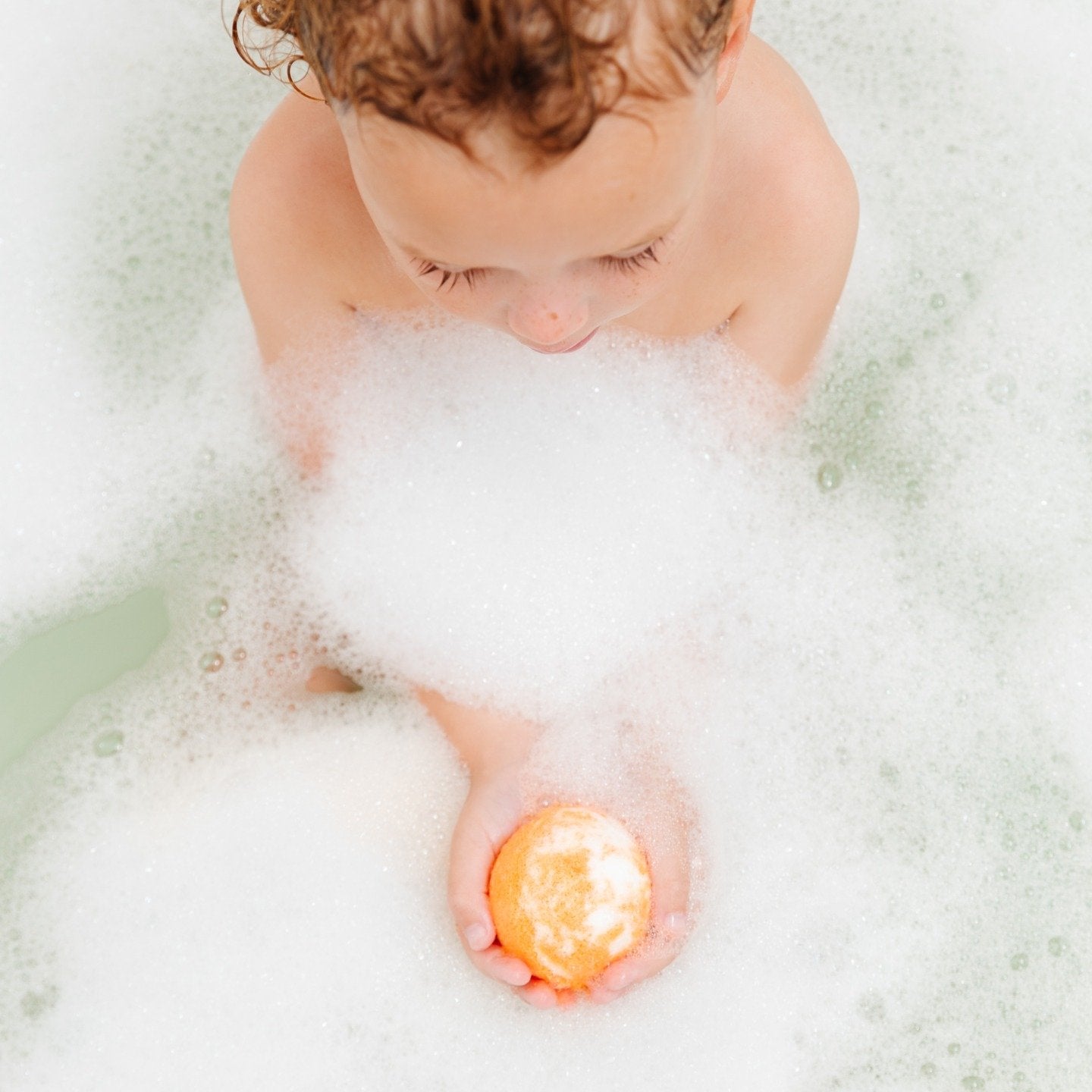 Child holding bath bomb in sudsy bath tub