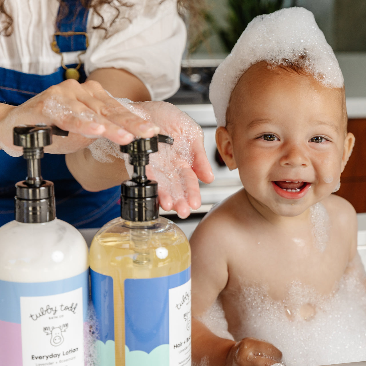 Smiling baby sitting at the sink with bubbles over his head and mother pumping wash from the side.