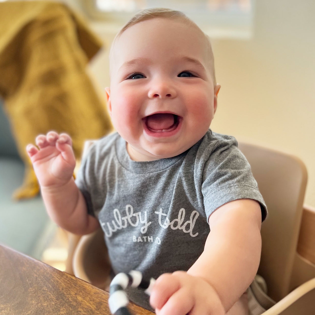Baby wearing Tubby Todd grey baby tshirt while sitting in high chair with big smile