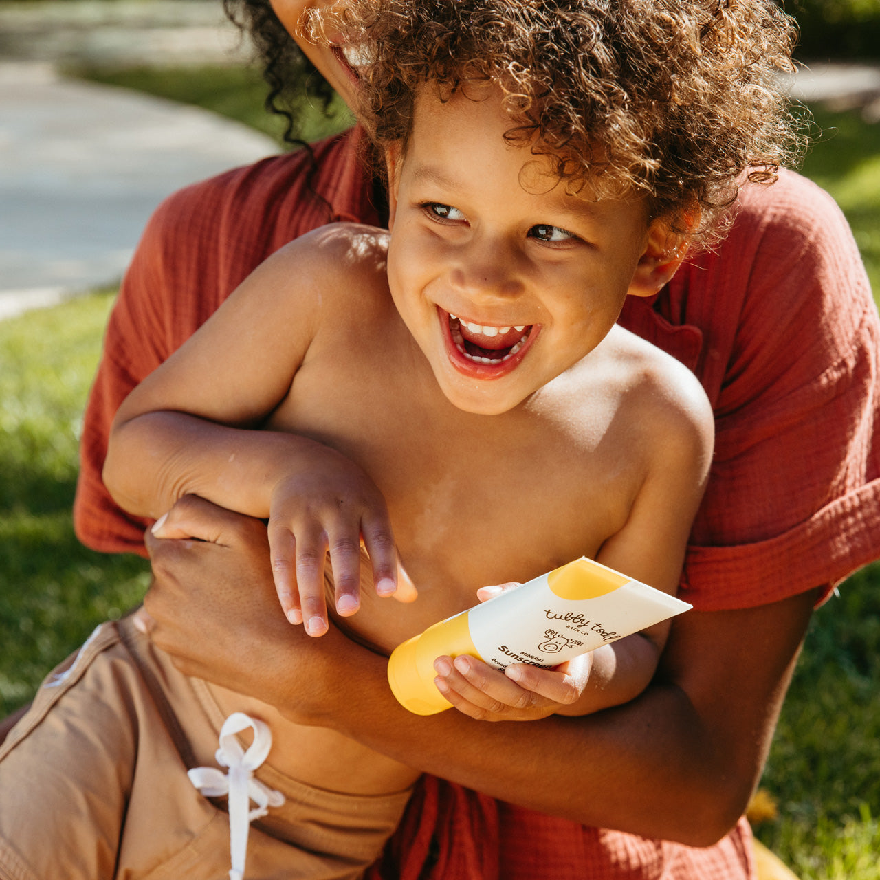 Mom holding laughing child on grass while holding sunscreen